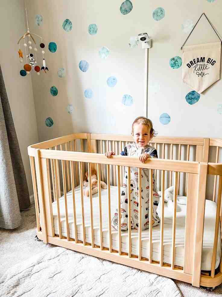 A wooden convertible crib and a baby standing inside it 