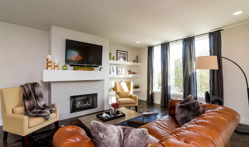 Floor to ceiling window with curtains and brown leather sofa with a TV above the fireplace