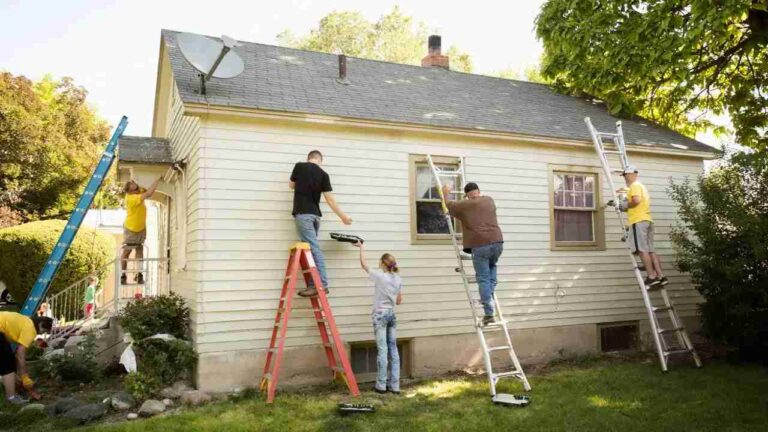 five-people-visible-around-the-house-multiple-ladder-are-in-the-use-people-were-positioned-at-various-heights-house-has-white-siding-and-roof