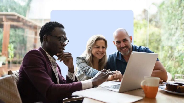 one-man-in-a-suit-is-pointing-at-a-laptop-screen-appearing-to-explain-something-a-woman-and-man-are-sitting-beside-him-smiling-and-looking-at-laptop