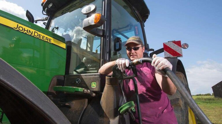 a-farmer-wearing-sunglasses-a-cap-and-a-maroon-shirt-and-filling-a-green-and-yellow-tractor-with-fuel