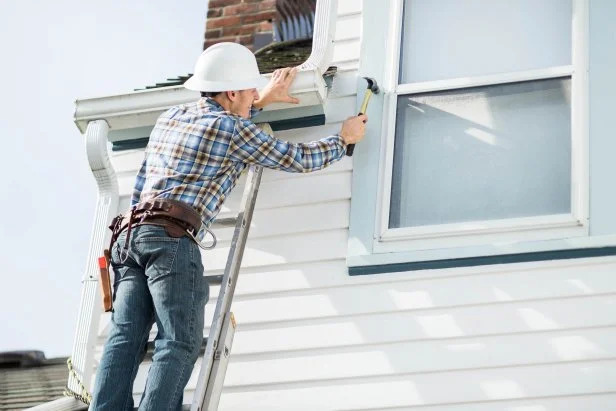How to Get Your Home Ready Before Selling a-man-is-standing-on-a-ladder-using-a-power-drill-to-work-on-the-white-siding-or-trim-of-a-house-exterior