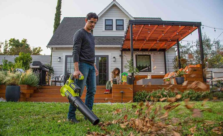Spring Home Prep Checklist: How to Prevent Pests Before Peak Season a-man-standing-on-a-lawn-operating-a-bright-lime-green-leaf-blower-to-clear-debris-and-there-is-white-house-in-a-background