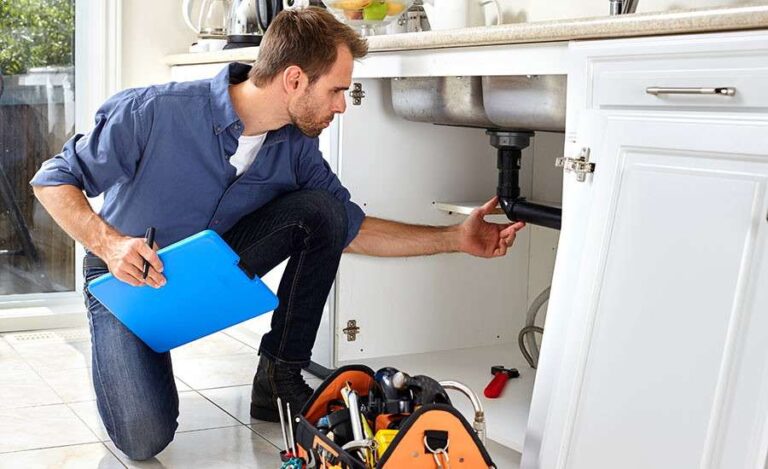 a-male-plumber-is-kneeling-down-holding-a-blue-clipboard-in-one-hand-while-reaching-under-the-sink-cabinet-with-other