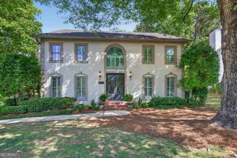 the-home-features-a-symmetrical-stucco-facade-with-an-arched-central-entryway-the-property-includes-lawn-mature-surroundings-trees