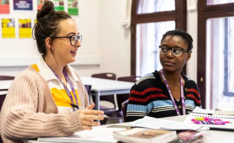 two-young-female-students-sitting-at-a-desk-inside-what-appears-to-be-a-classroom-setting